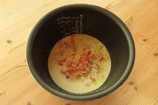 米と牛乳、水、調味料の入った炊飯器の内釜にカニカマと玉ねぎ、にんにくを入れる。